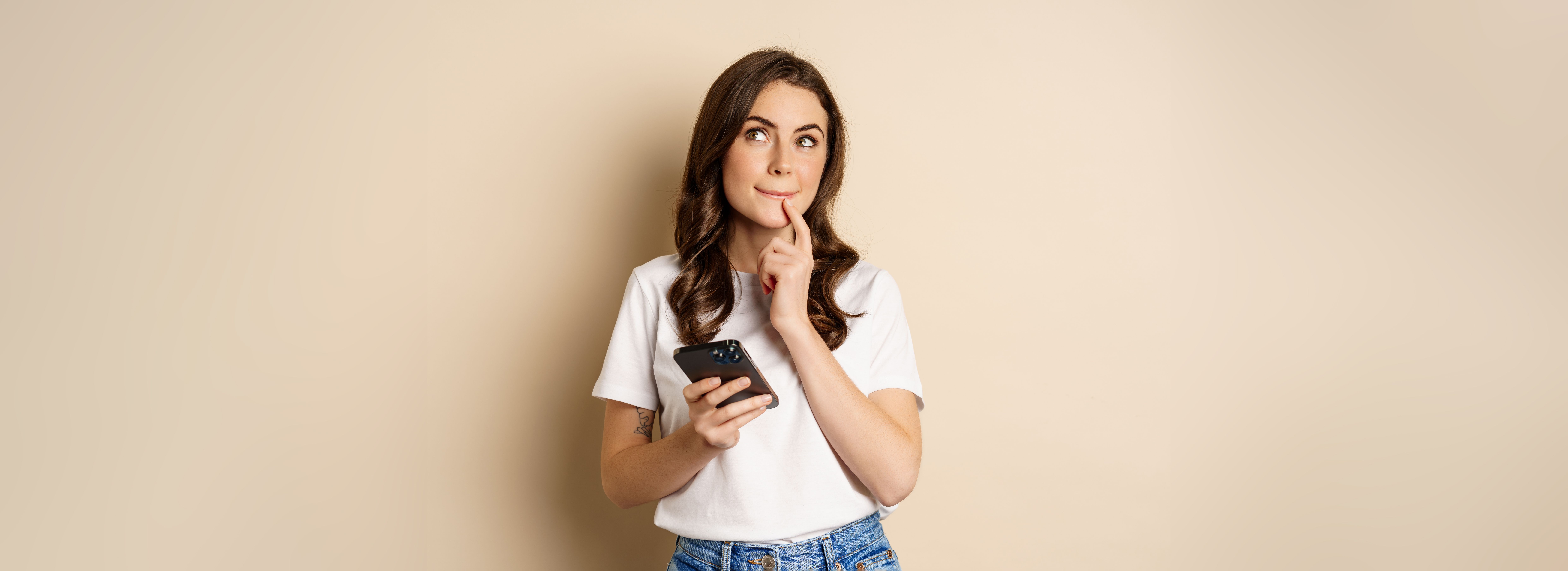 girl-thinks-holds-mobile-phone-looks-up-thoughtful-stands-beige-background.jpg