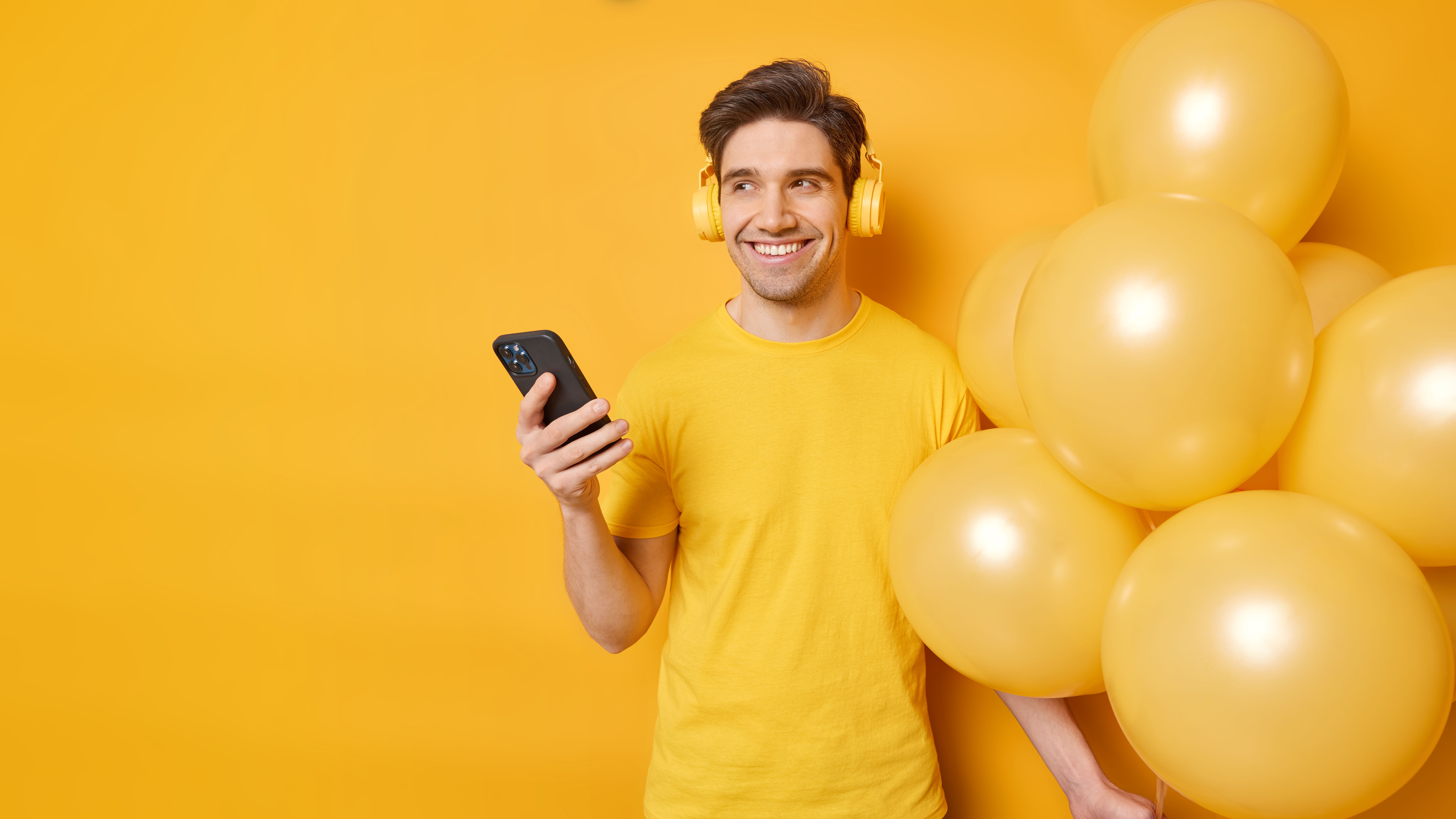 happy-dreamy-man-looks-away-with-cheerful-expression-listens-smartphone-andstereo-headphones-listening-music-holds-bunch-balloons-isolated-yellow-background-copy-space-your-promotion.jpg