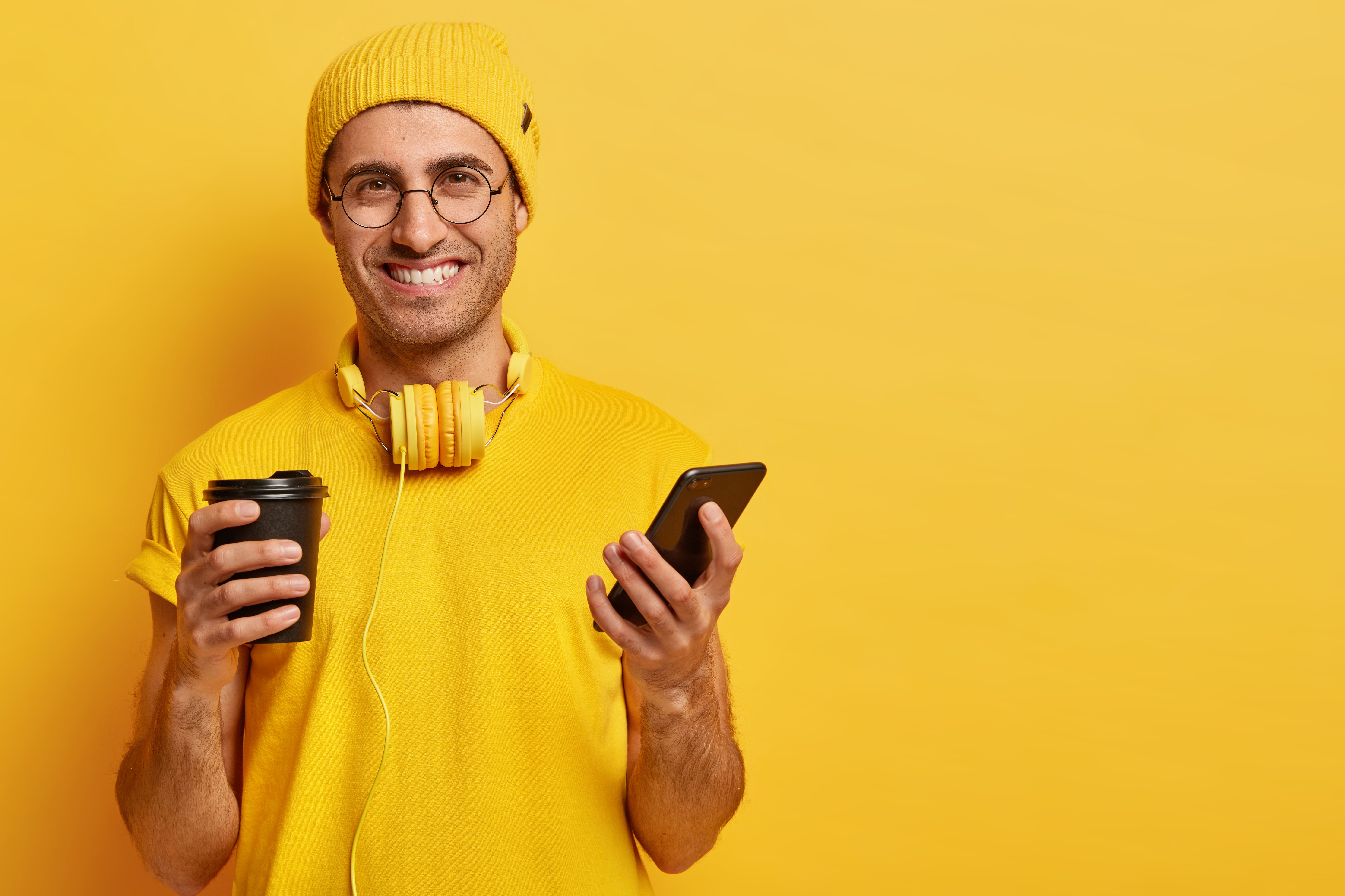 portrait-handsome-cheerful-young-man-with-pleased-face-expression-holds-mobile-phone-sends-text-messages-friends-drinks-takeaway-coffee-wears-spectacles-yellow-outfit-with-headphones.jpg