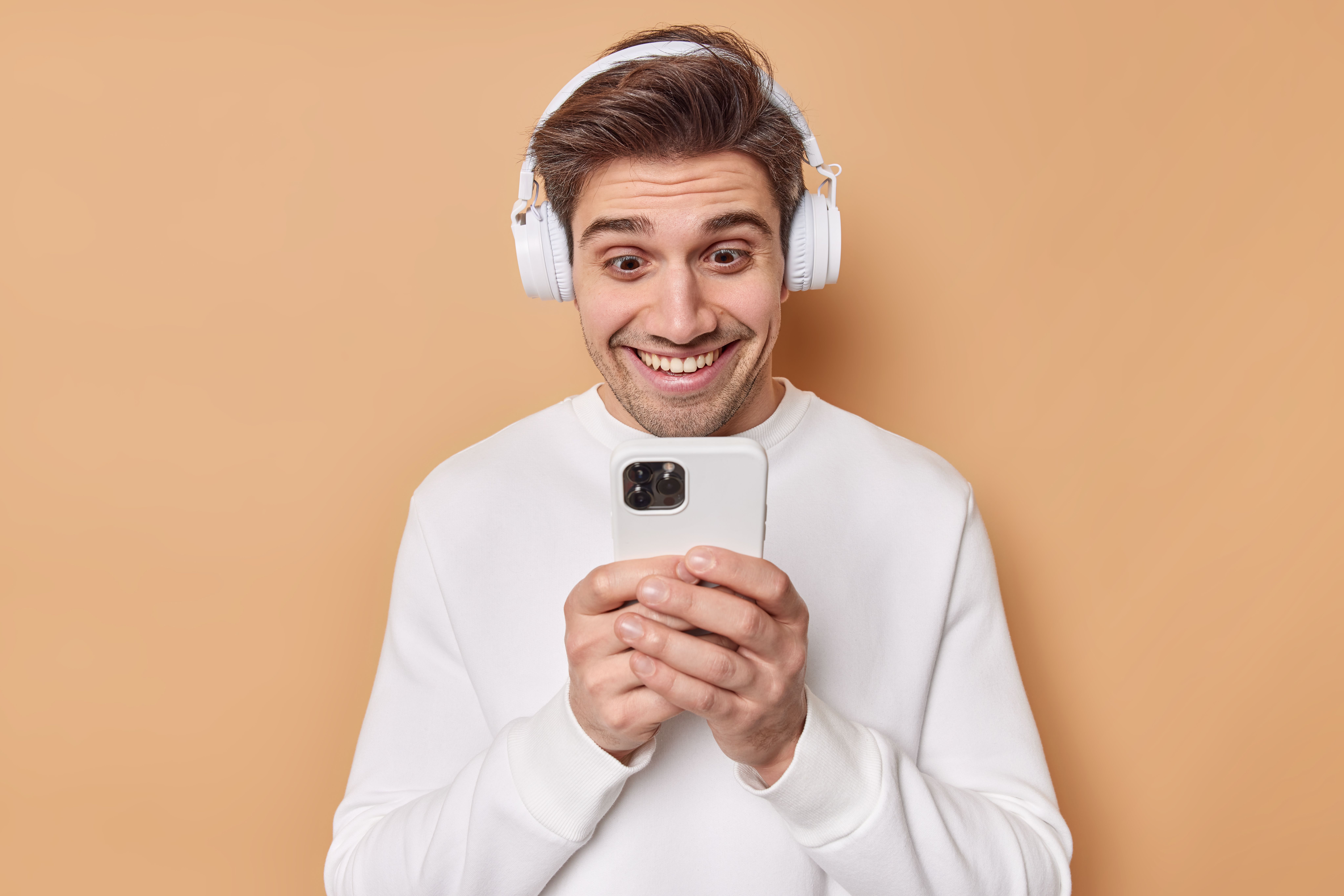 positive-young-man-looks-happily-smartphone-screen-reads-funny-story-leaves-comments-post-listens-audio-track-via-wireless-headphones-dressed-casual-jumper-isolated-beige-background.jpg