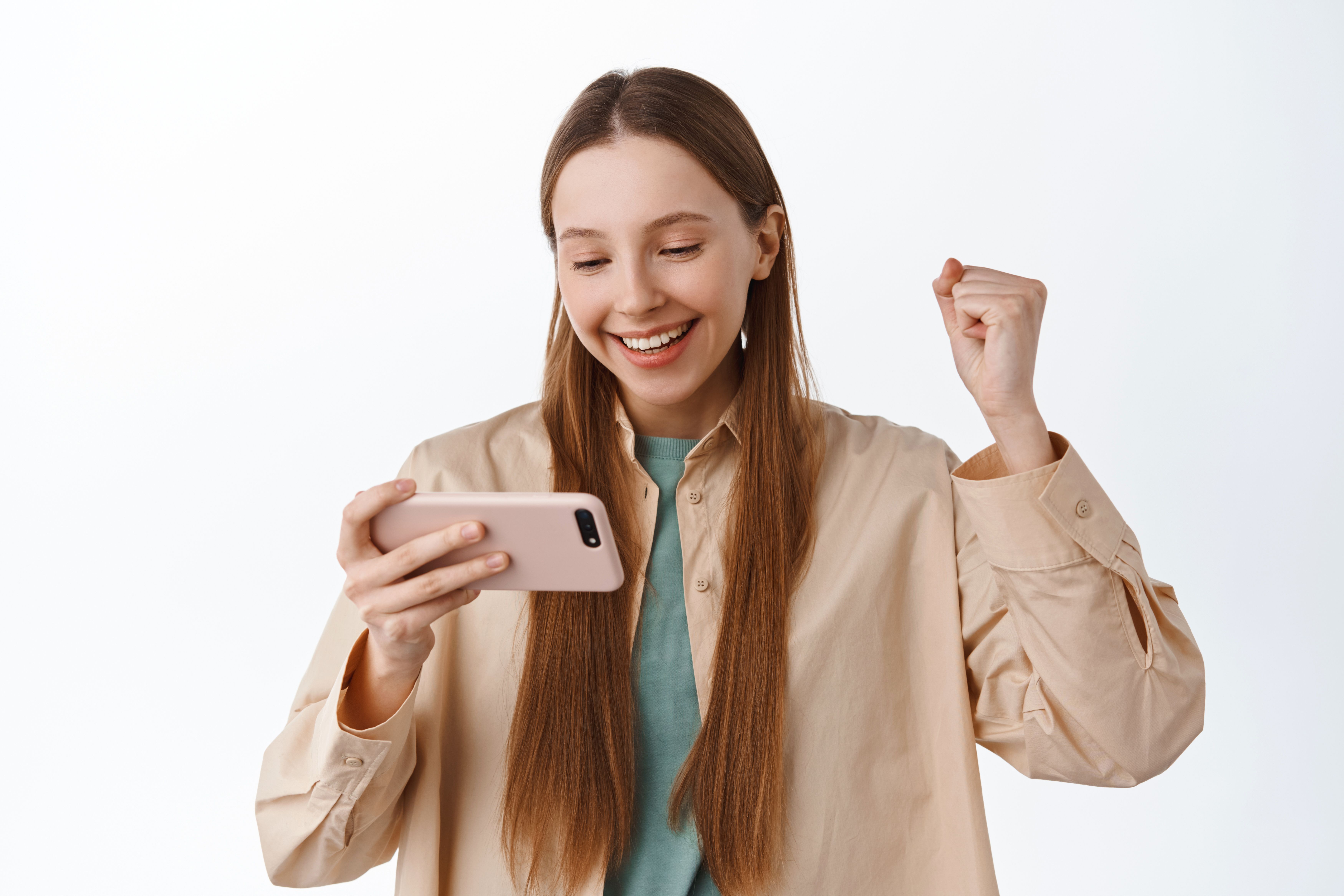 smiling-girl-looks-smartphone-rejoice-fist-pump-pleased-winning-mobile-video-game-celebrating-online-victory-achieve-goal-app-standing-white-wall.jpg