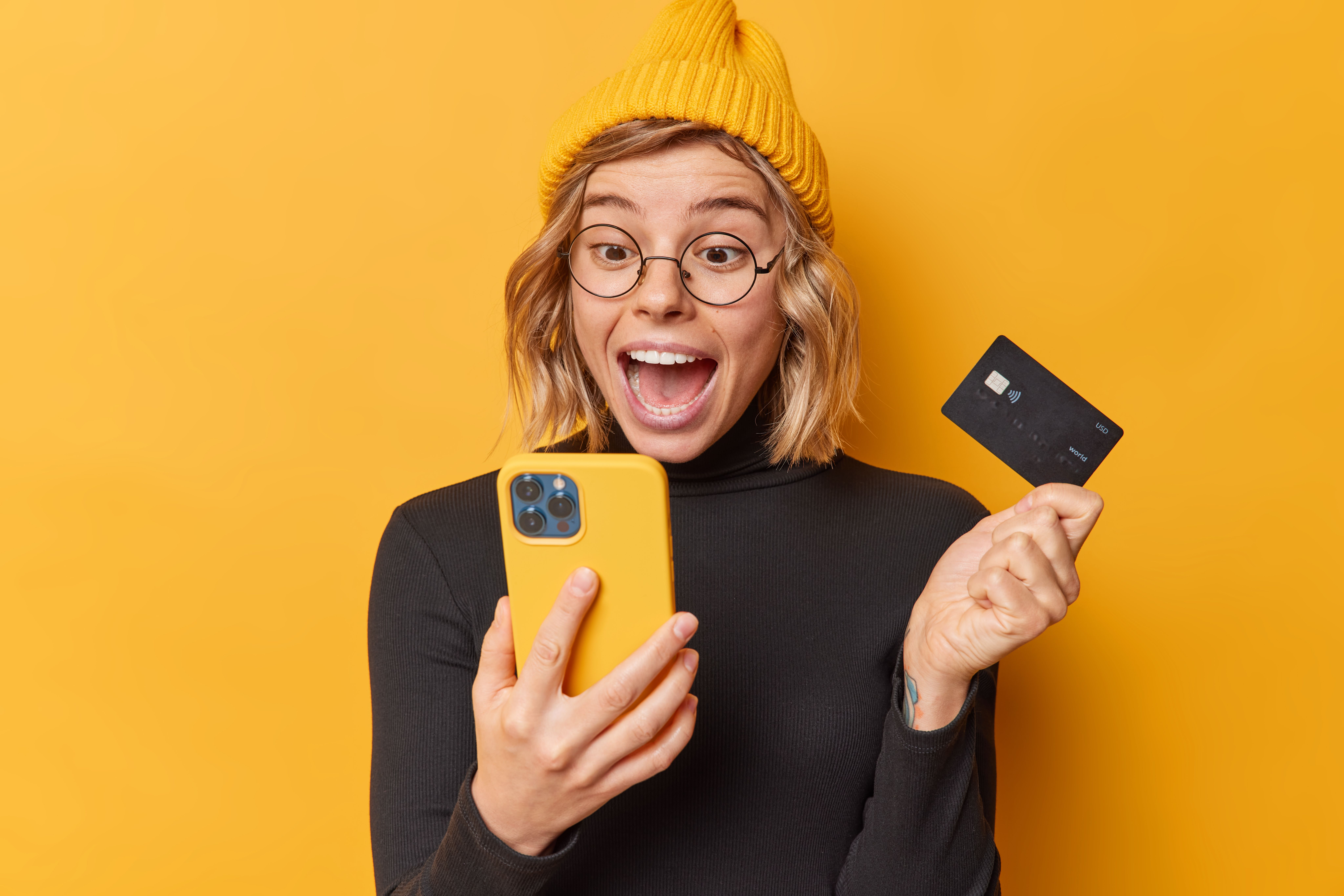 surpised-happy-woman-wears-hat-casual-black-turtleneck-looks-smartphone-app-screen-holds-credit-card-uses-bank-account-paying-online-isolated-yellow-background-gets-cashback.jpg